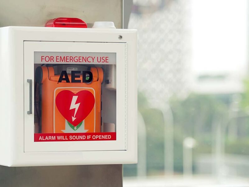 Picture showing an orange coloured defibrillator in a white coloured wall-mounted cabinet.