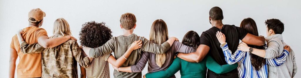 A diverse group of people seen from behind, standing side-by-side with their arms around each other's shoulders against a plain white background.