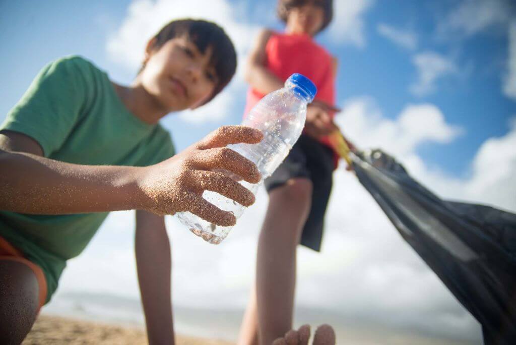 A close-up, low-angle shot of a child picking up a sandy plastic water bottle on a beach during a cleanup.
