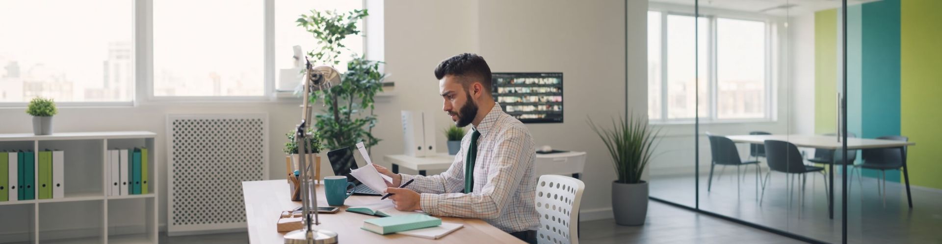 A male trainer sitting at a modern office desk reviewing training assessment paperwork with a pen and laptop.