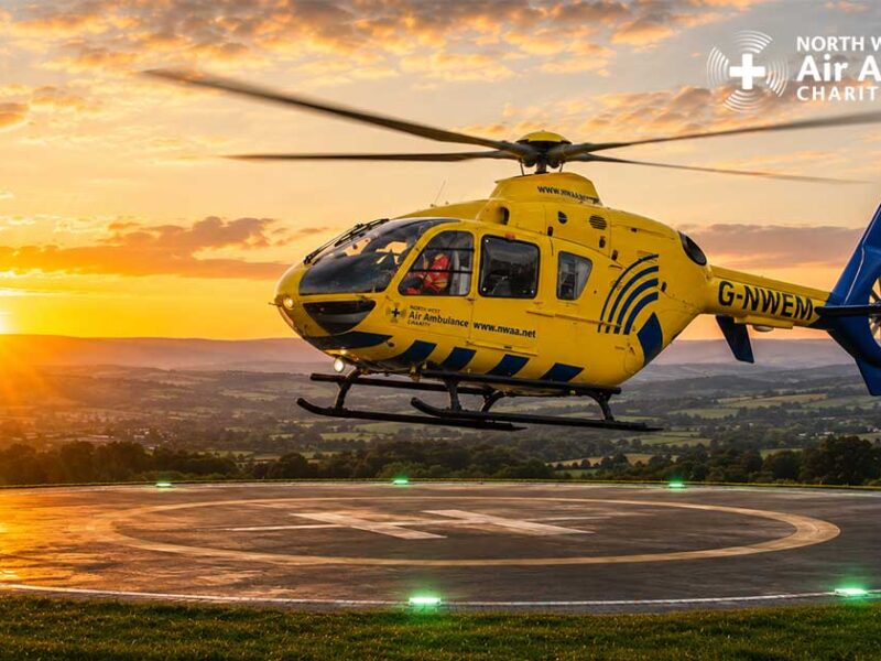 A yellow and blue North West Air Ambulance Charity helicopter stationed on a hillside helipad at sunset.