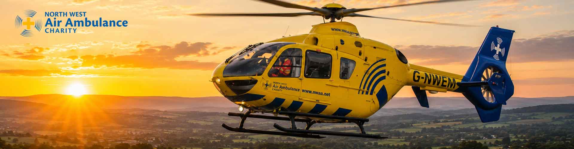 A yellow and blue North West Air Ambulance Charity helicopter stationed on a hillside helipad at sunset.