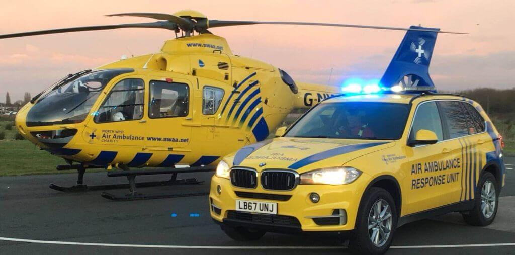A bright yellow and blue North West Air Ambulance helicopter parked next to a matching BMW emergency response SUV with flashing blue lights at dusk.