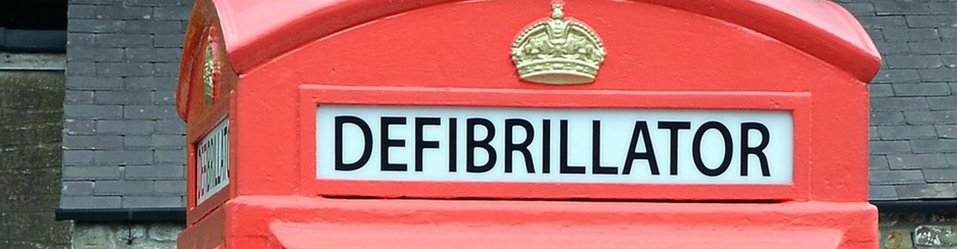 A close-up of a traditional red British telephone box repurposed as a community public access defibrillator station, showing clear black 'DEFIBRILLATOR' signage and the gold crown emblem against a slate roof background.