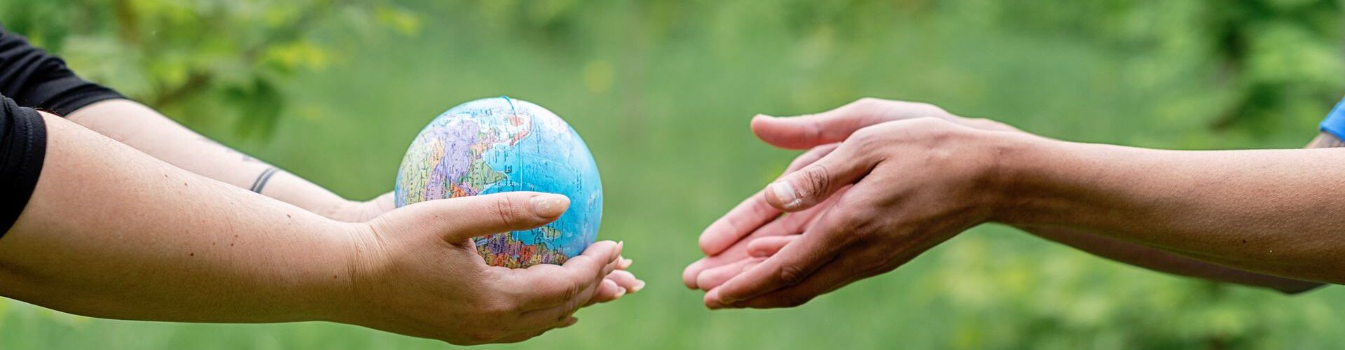One person gently holding a small model of the Earth and passing it into another person's open hands, set against a soft, out-of-focus green foliage background.